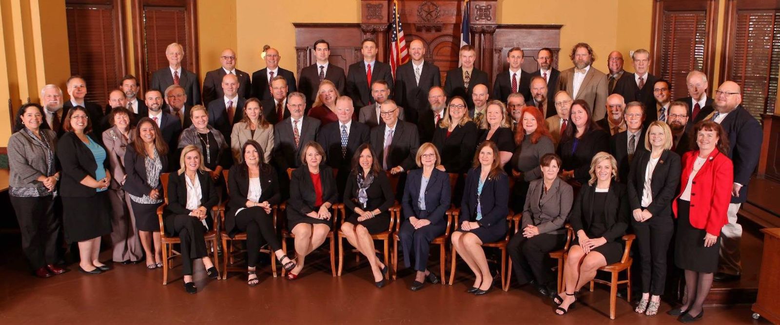 Group portrait of the members of the Ellis County Bar Association, gathered before the judge's bench in a historic Ellis County courtroom, with the United States and Texas flags behind them.